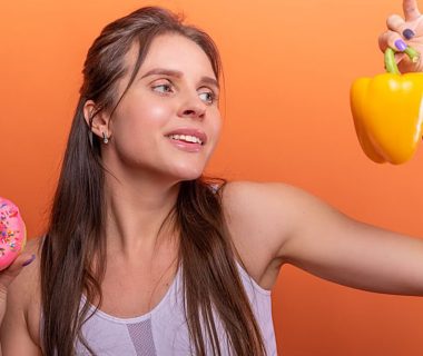 Woman in sports bra juggling choice between a donut and bell pepper on orange background.