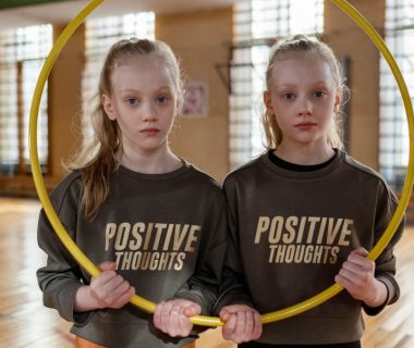 Twin girls in gym holding a hula hoop, wearing 'Positive Thoughts' sweatshirts.
